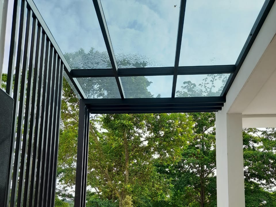 A lean-to glass roof on a slim black frame over a side terrace, looking out past a potted shrub to mature trees.